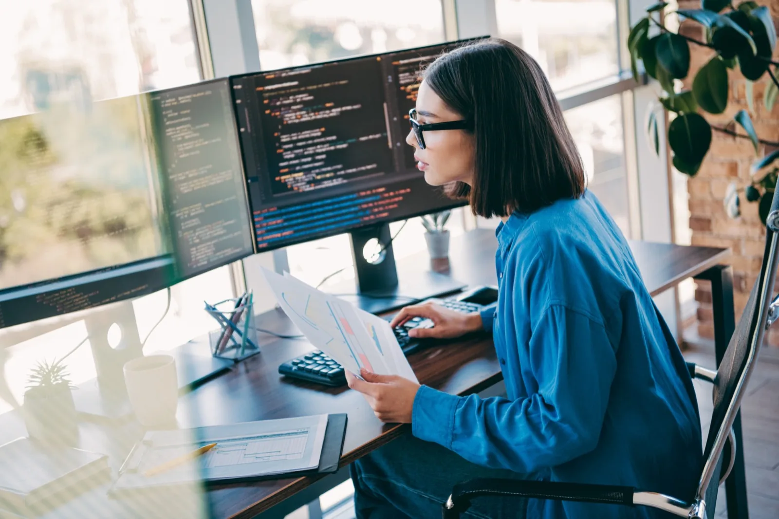 Engineer in glasses reviewing printed charts beside two monitors of infrastructure code